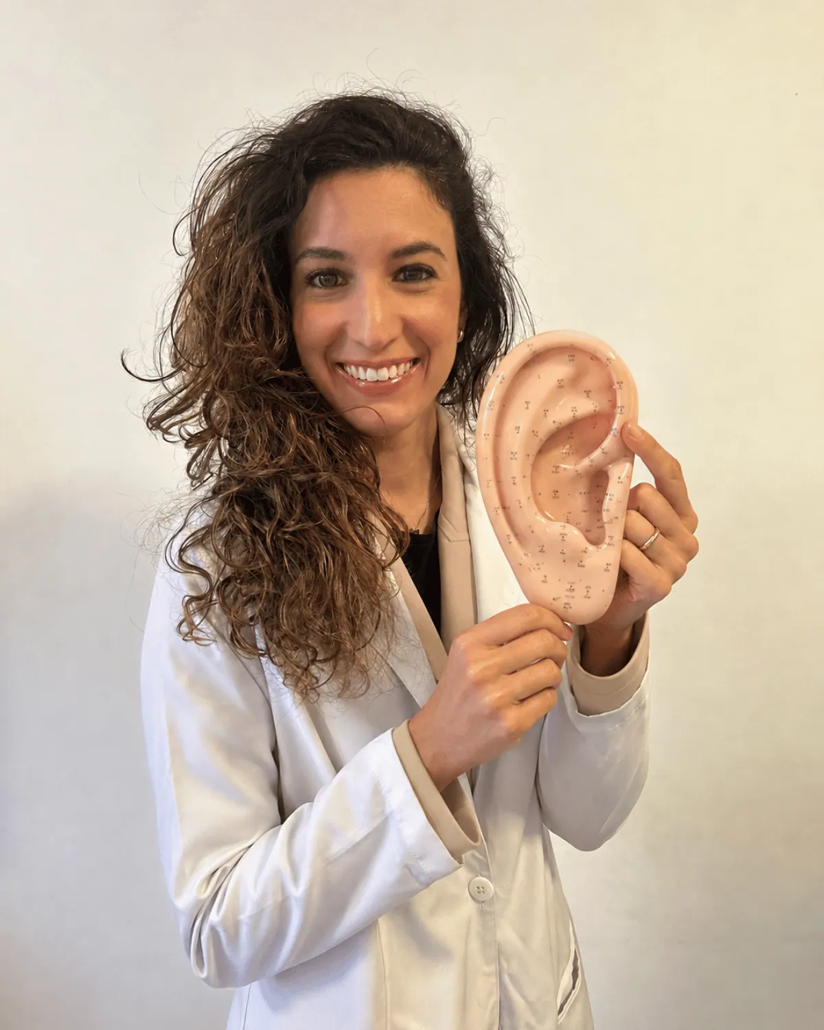 Fernanda Pradines sorrindo, segurando uma orelha anatômica com os pontos auriculares marcados
